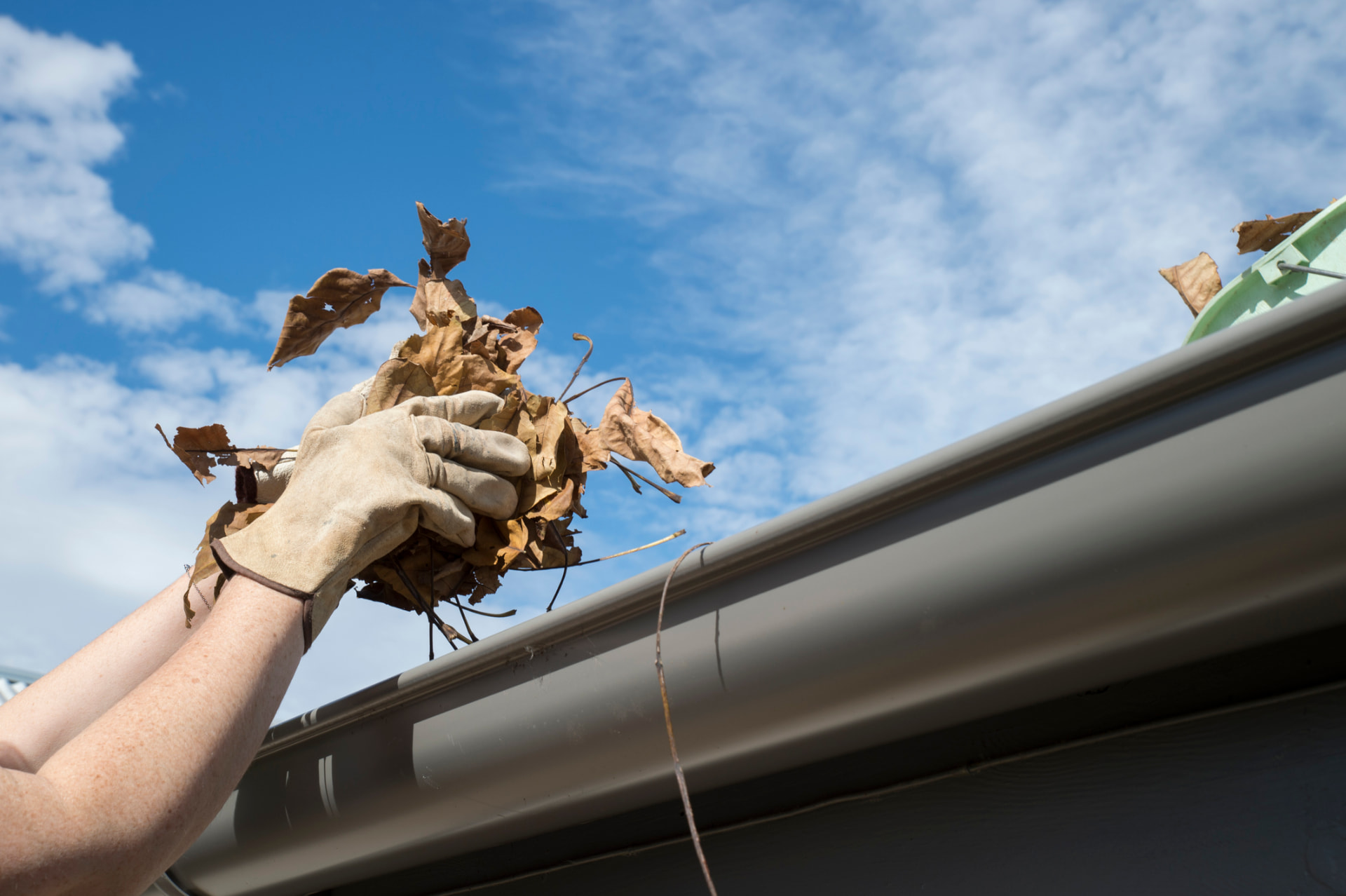 Gloved hand lifting a clump of dry leaves out of a home gutter against a blue sky