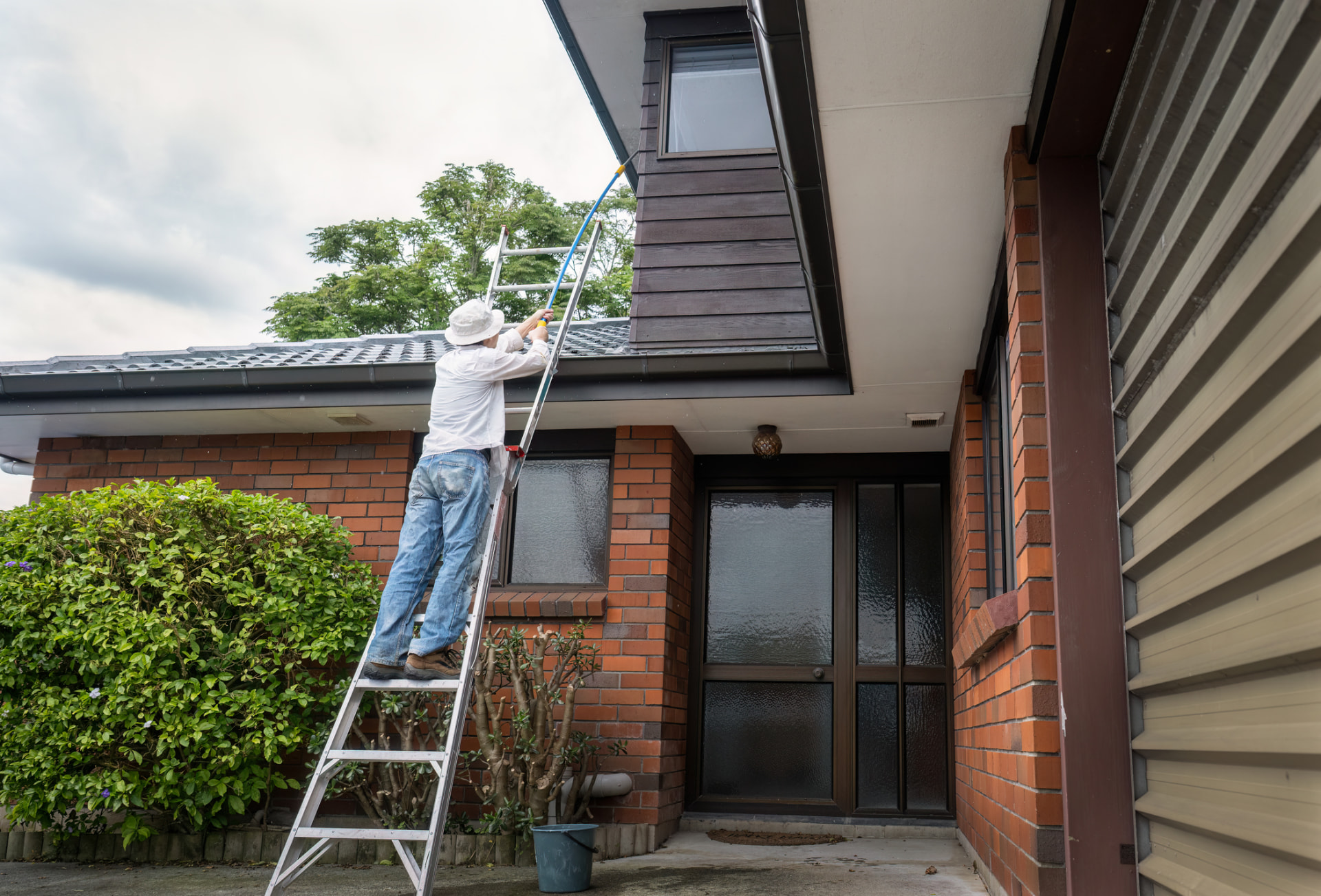 Technician on a ladder cleaning upper-story windows and eaves of a brick home